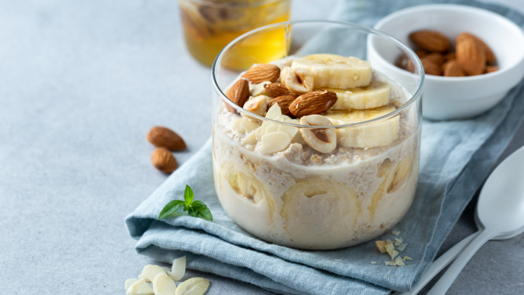 Glass jar of instant oats topped with banana slices, almonds, and honey, prepared by a trusted instant oats manufacturer, served on a blue napkin with extra nuts on the side.